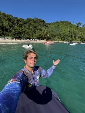 Cairê dentro do barco, em frente a uma enseada com barcos ancorados e a mata atrás.