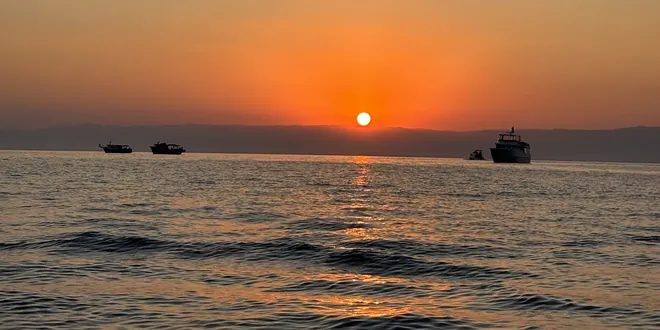 O sol nascendo sobre o mar, com barcos ancorados recortados contra a luz.