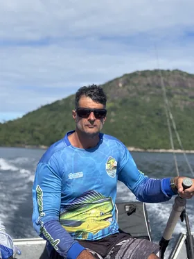 Leandro ao leme do barco, de camisa de pesca, com o costão verde ao fundo.
