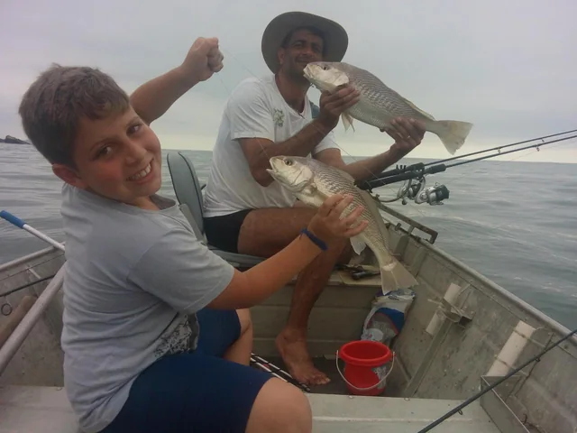 Foto antiga de pescaria: um homem e um menino em pé num barco de alumínio, cada um segurando um peixe.