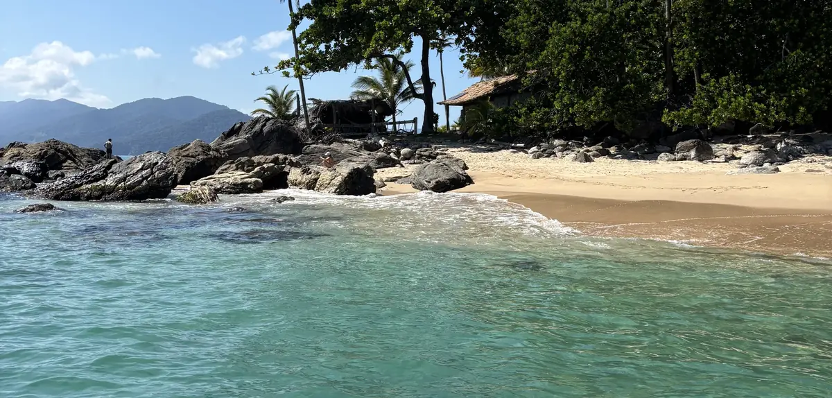 A praia vista de dentro da água transparente, com coqueiros, um quiosque de telhado de sapé e o costão de pedras na beira.