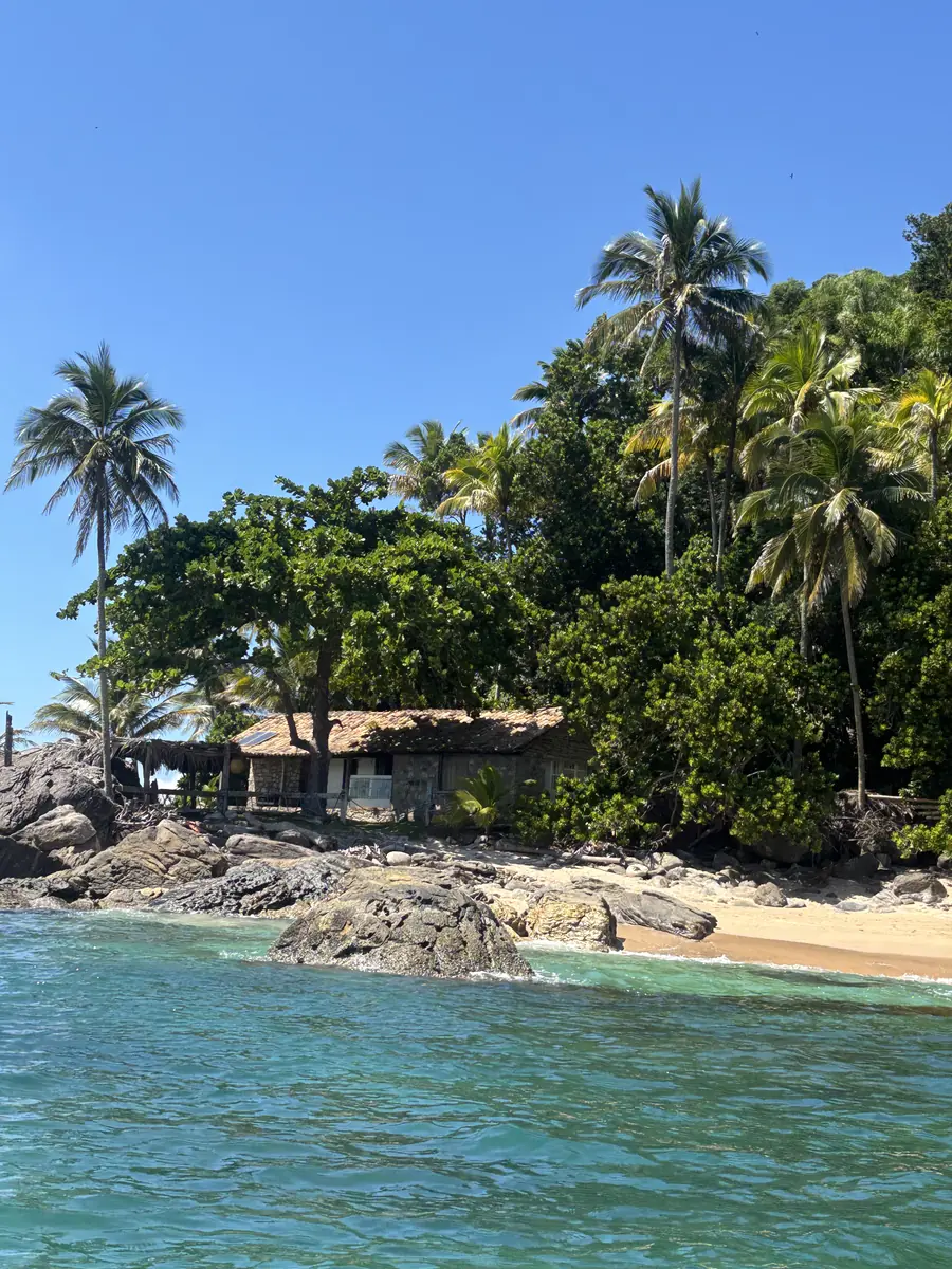 Casa de telhado de sapê na beira da praia da Ilha dos Gatos, entre coqueiros e pedras.