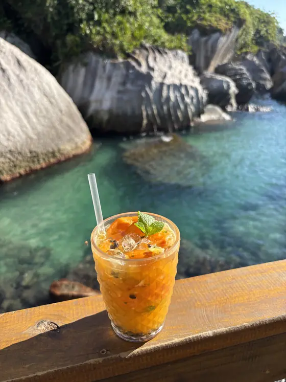Caipirinha apoiada na mureta do quiosque, com a piscina natural do Montão do Trigo ao fundo.