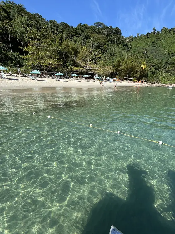 Água rasa e transparente na chegada à praia, com a estrutura ao fundo.